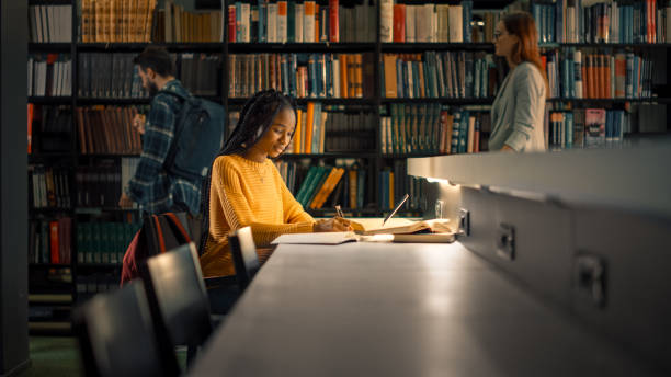 Studentss University Library: Gifted Black Girl uses Laptop, Writes Notes for the Paper, Essay, Study for Class Assignment. Students Learning, Studying for Exams College. Side View Portrait with Bookshelves